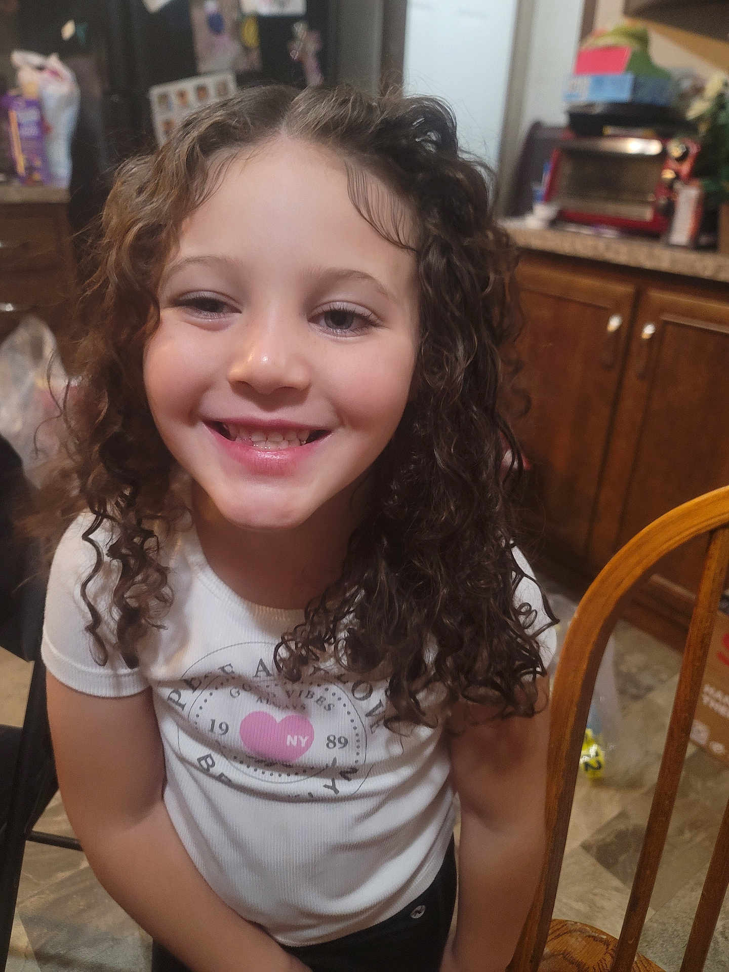 A'Mariyana is registered to the contest to win money with this photo: child, girl, smile, curly_hair, white_shirt, pink_heart, kitchen, wooden_chair, cabinets, indoor, happy, person, portrait, casual_clothing, young, seated, home, face, expression