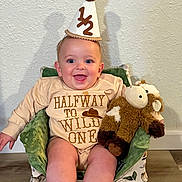 Cyle is registered to the contest to win money with this photo: baby, party_hat, smiling, plush_toy, child, indoor, cushion, chair, clothing, text_on_clothing, celebration, happy, person, portrait, infant, decor, floor, wall, seated, cute