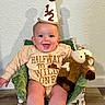 baby, party_hat, smiling, plush_toy, child, indoor, cushion, chair, clothing, text_on_clothing, celebration, happy, person, portrait, infant, decor, floor, wall, seated, cute