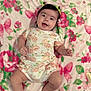 baby, infant, child, smiling, lying_down, floral_pattern, blanket, onesie, hair_ties, cute, happy, skin, face, hands, legs, feet, indoors, soft_texture, colorful, comfort