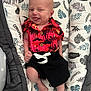 baby, infant, smiling, clothing, red_shirt, black_shorts, cushion, leaf_pattern, happy, face, skin, tongue_out, relaxed, indoors, cute, person, young_child, lying_down, fabric, comfortable