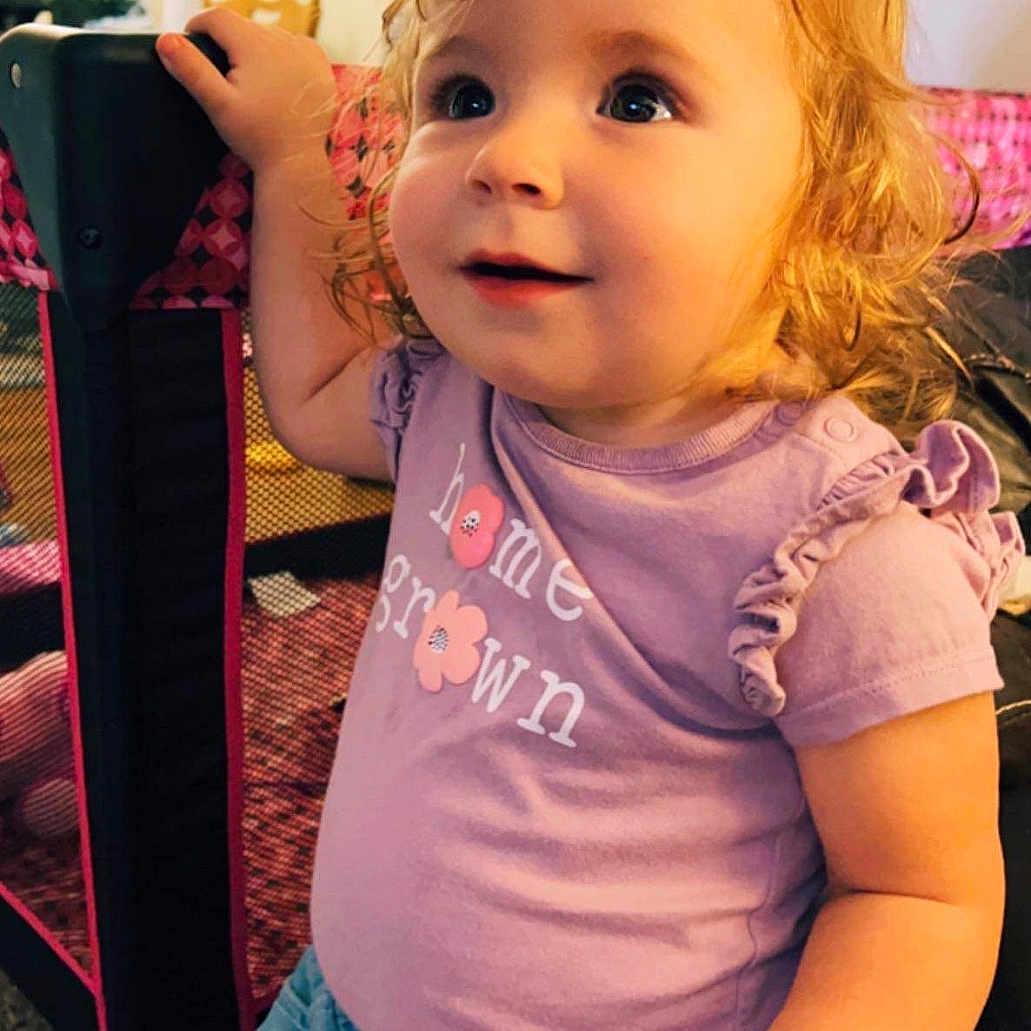 Journeigh is registered to the contest to win money with this photo: bright_eyes, carpet, casual_clothing, child, curious, curly_hair, cute, denim_shorts, hand, happy, home_interior, indoors, person, playpen, portrait, purple_shirt, seated, smiling, toddler, young_child