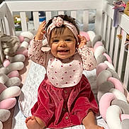 Amaya is registered to the contest to win money with this photo: baby, child, smile, crib, headband, bib, dress, corduroy, pink, red, pastel, braided_cushion, soft_toy, happy, sitting, indoors, cute, infant, portrait, playful