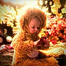 bokeh, brown_hair, child, costume, cozy, cute, face, festive, flowers, furry_outfit, holding_object, holiday_lights, indoor, photography, portrait, red_object, sitting, soft_focus, toddler, warm_lighting