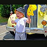 child, chrome, front_grille, gray_pants, happy, hat, headlight, outdoors, playful, portrait, road, smiling, sunlight, toddler, toy, trees, vehicle, vintage_truck, white_shirt, yellow_vehicle