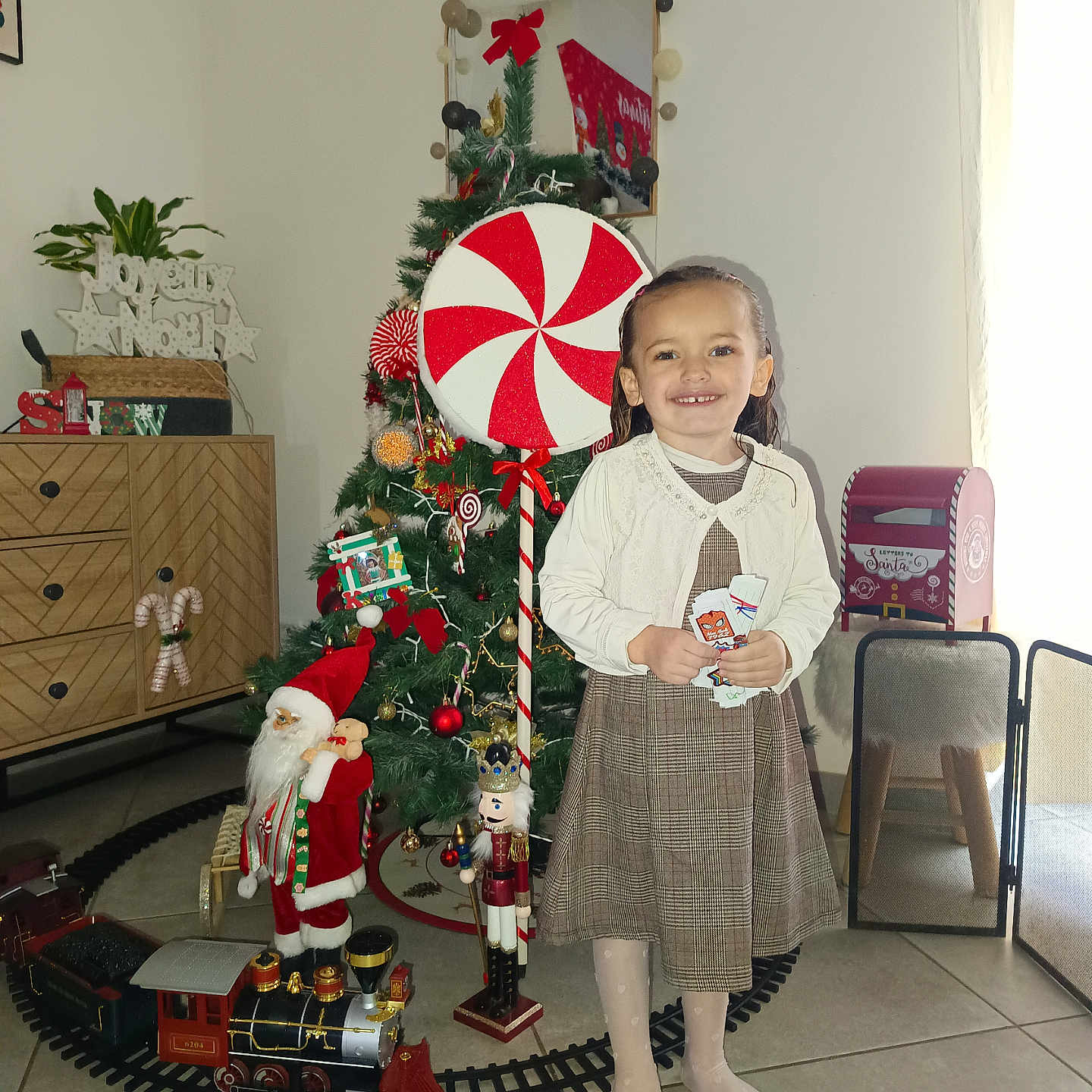Kataleya participe au concours pour gagner de l'argent avec cette photo : cabinet, cardboard_letters, child, christmas_tree, dress, festive, floor_tiles, furniture, girl, holiday_decorations, indoor, mailbox, mirror, nutcracker, peppermint_decoration, santa_figurine, smiling, tights, toy_train, window