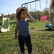 Kayden a rejoint le concours — aidez-le/la à gagner de superbes lots ! black_pants, blue_tank_top, casual_clothing, child, curly_hair, daylight, grass, hat, nature, outdoor, park, person, playground, slide, sneakers, sunny, swings, thinking, toddler, young_child