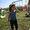 black_pants, blue_tank_top, casual_clothing, child, curly_hair, daylight, grass, hat, nature, outdoor, park, person, playground, slide, sneakers, sunny, swings, thinking, toddler, young_child