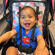 Ava joined the competition — help win amazing prizes! baby, blue_shirt, child, clothing, colorful, cute, geometric_pattern, hands, happy, indoor, person, playful, portrait, seat, sitting, smiling, strawberry_graphic, stroller, toddler, young_child