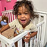 toddler, child, crib, white_crib, curly_hair, smiling, baby, indoor, pink_wall, teddy_bear, stuffed_animal, clothing, person, happy, playful, nursery, bedroom, furniture, cute, portrait