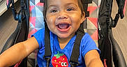 Ava is registered to the contest to win money with this photo: baby, black_stroller, blue_shirt, cheerful, child, colorful_pattern, cute, hands, happy, holding, indoor, person, playful, portrait, seatbelt, smile, strawberry_graphic, stroller, toddler, young_child