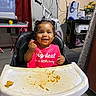 toddler, child, high_chair, messy, food, bib, smiling, indoor, bicycle, table, chair, curtain, floor, headphones, printer, flowers, toy, person, happy, home