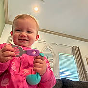 Ivy is registered to the contest to win money with this photo: toddler, child, smiling, pink_clothing, flamingo_pattern, toy, kitchen_utensils, indoor, living_room, curtains, window, sofa, ceiling_light, mirror, happy, person, playful, cute, home, casual