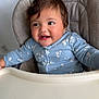 baby, child, high_chair, blue_clothing, floral_pattern, smiling, happy, indoor, face, person, young_child, cute, kitchen, utensil_pattern, seat, infant, table, portrait, sitting, cheerful