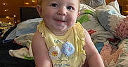 Athena is registered to the contest to win money with this photo: baby, child, infant, smiling, portrait, sitting, indoors, bedroom, bed, blankets, pillows, fairy_lights, warm_lighting, yellow_outfit, floral_clothing, barefoot, blue_eyes, happy, cozy, photography