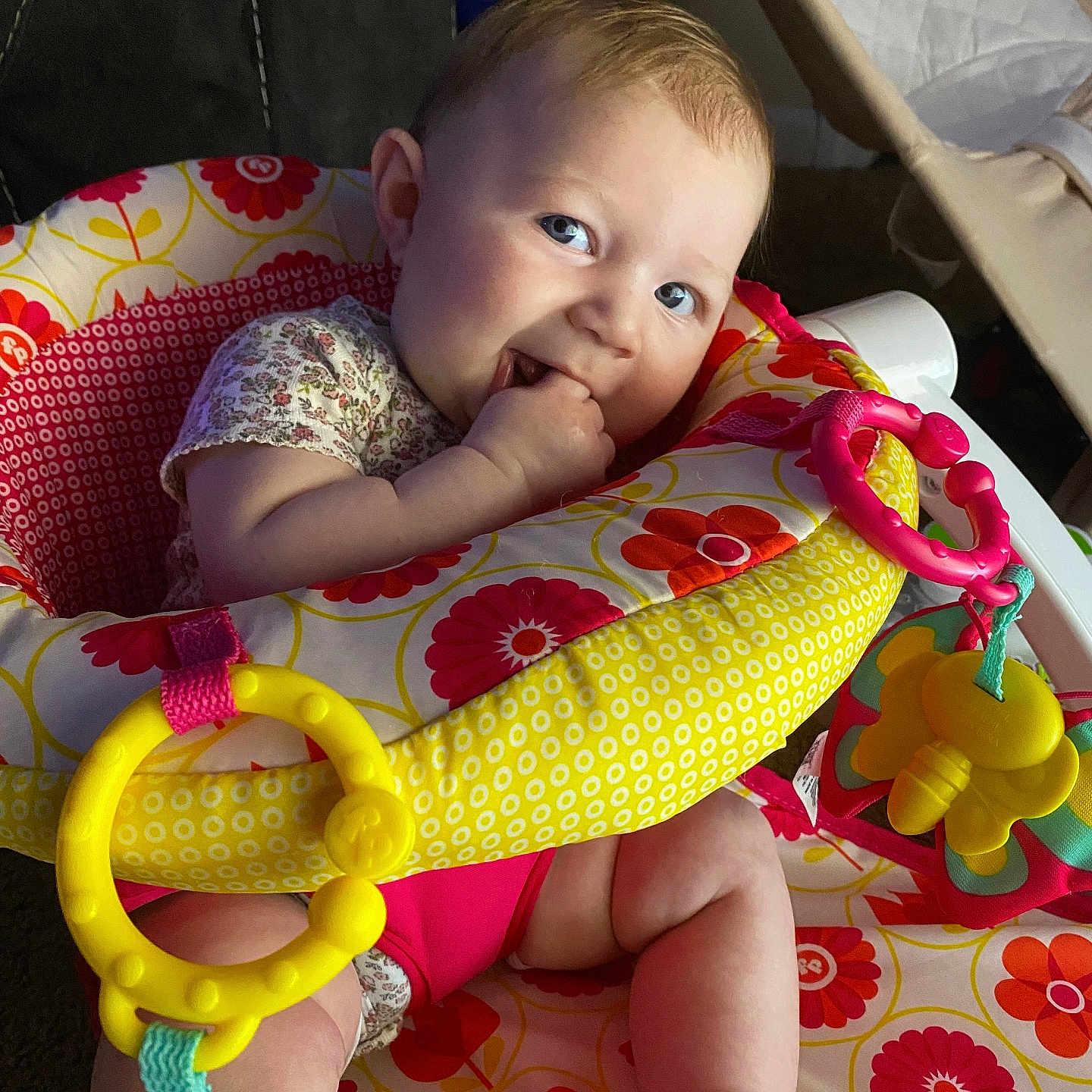 Athena is registered to the contest to win money with this photo: baby, child, infant, smiling, sitting, activity_center, baby_toy, teething, hand, blue_eyes, floral_pattern, colorful, indoor, portrait, playtime, jumper, sock, pink_clothing, yellow_toy, happy