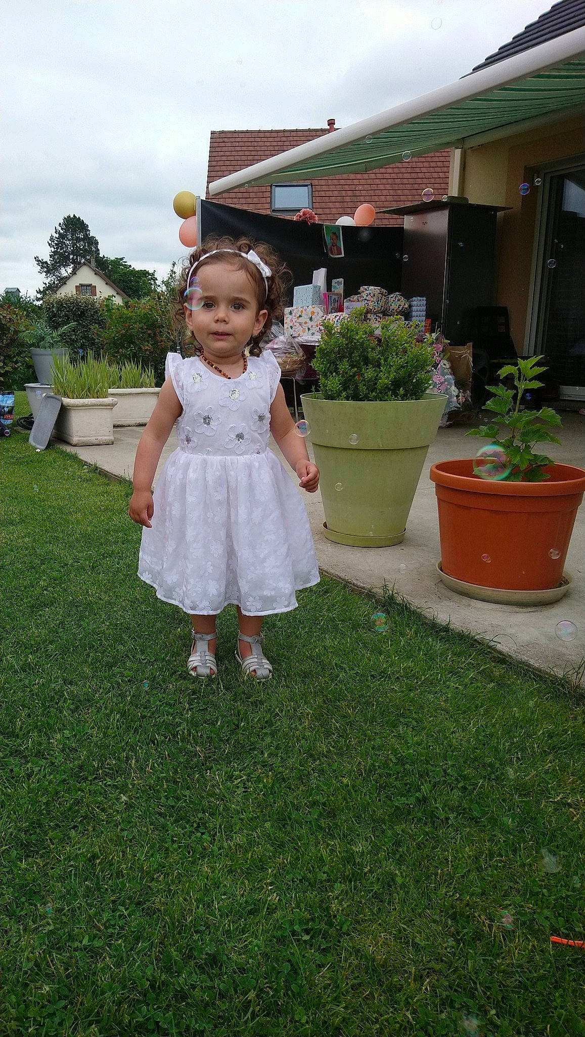 Noély participe au concours pour gagner de l'argent avec cette photo : bridal_clothing, day_dress, dress, event, flooring, flower, flowerpot, grass, happy, house, houseplant, landscape, one_piece_garment, person, petal, plant, shrub, sky, summer, toddler