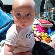 Atley is registered to the contest to win money with this photo: baby, infant, child, onesie, chubby_cheeks, big_eyes, sitting, toy, colorful_toy, blanket, indoor, cluttered_background, shelf, door, bottle, person, portrait, candid, skin, gaze