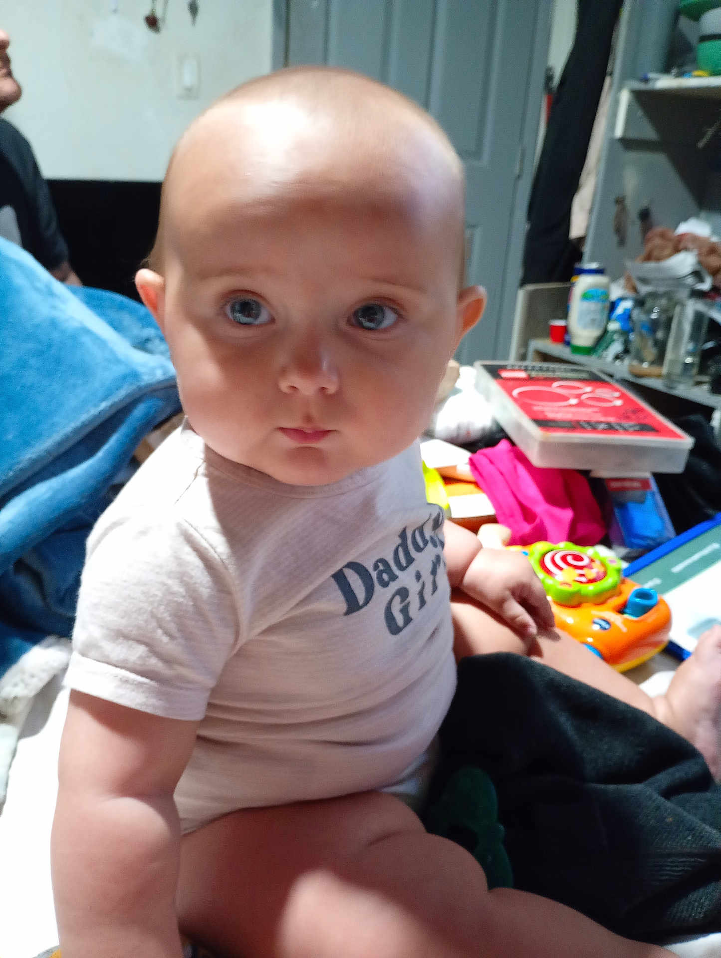 Atley is registered to the contest to win money with this photo: baby, infant, child, onesie, chubby_cheeks, big_eyes, sitting, toy, colorful_toy, blanket, indoor, cluttered_background, shelf, door, bottle, person, portrait, candid, skin, gaze