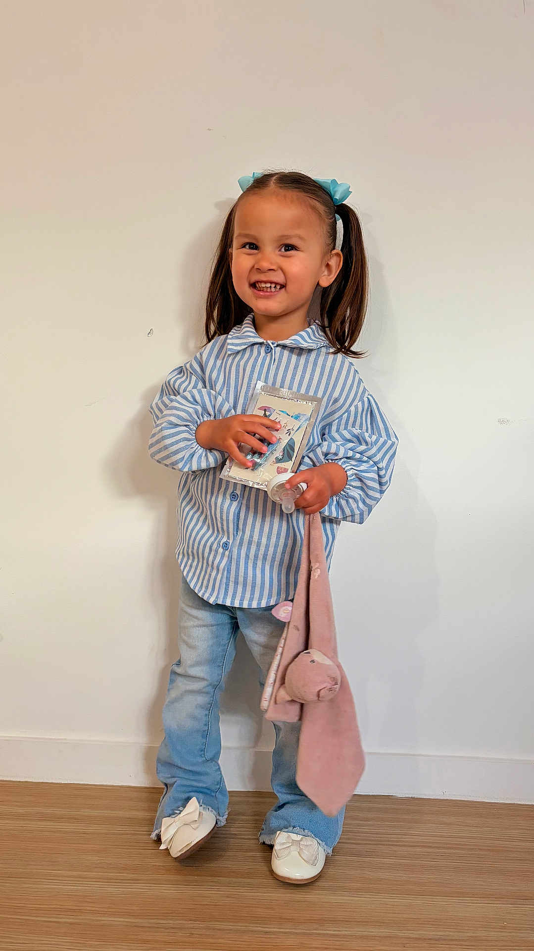 Layana a rejoint le concours — aidez-le/la à gagner de superbes lots ! child, girl, pigtails, blue_bows, striped_shirt, jeans, white_shoes, smile, standing, indoor, plush_toy, book, happy, cute, person, portrait, young_child, floor, wooden_floor, wall