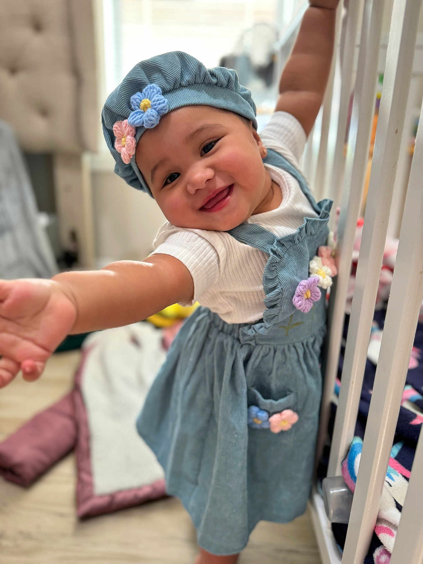 Marcela joined the competition — help win amazing prizes! baby, child, smiling, blue_dress, flower, hat, crib, blanket, indoor, happy, person, cute, toddler, playful, clothing, furniture, soft_toys, floor, home, portrait