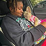 person, dog, puppy, sleeping, car_interior, seat, clothing, dreadlocks, black_hoodie, pink_outfit, window, outdoor, tree, nature, affection, holding, closeup, portrait, relaxed, cute