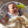 baby, infant, blanket, mittens, baby_seat, tropical_design, tongue_out, happy, indoors, wood_floor, playful, child, cute, person, soft_lighting, smiling, young, seated, clothing, fabric