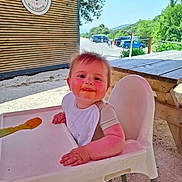 Léo participe au concours pour gagner de l'argent avec cette photo : baby, bib, car, child, daylight, face, feeding, food, greenery, happy, high_chair, outdoor, person, picnic_table, road, sign, smiling, spoon, sunny, wooden_wall