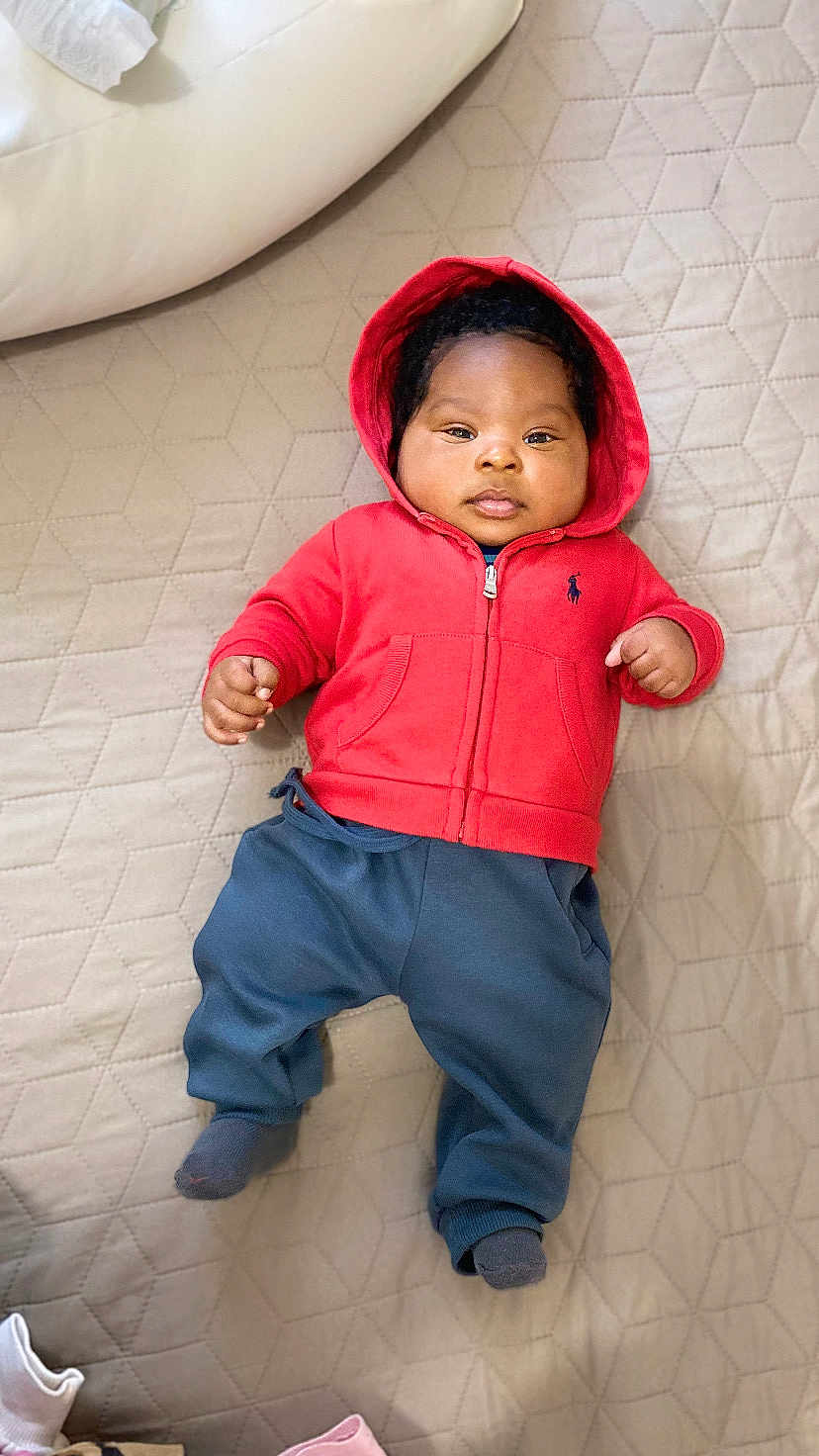 Maël participe au concours pour gagner de l'argent avec cette photo : baby, infant, child, hoodie, red_clothing, blue_pants, socks, lying_down, beige_quilt, quilted_bedspread, face, hands, feet, indoors, cute, portrait, sleepwear, comfortable, soft_lighting, casual