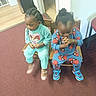 child, toddler, two_children, sitting, chairs, indoor, carpet, wooden_chair, blue_clothing, turquoise_clothing, princess_design, spiderman_design, sneakers, braided_hair, curly_hair, hands_clasped, flooring, wall, playful, cute