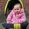 child, girl, stroller, pink_jacket, winter_clothing, outdoor, brick_wall, puffy_jacket, tights, cold_weather, seated, person, baby_stroller, portrait, expression, gloves, urban, young_child, casual, cute
