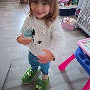 Angelina participe au concours pour gagner de l'argent avec cette photo : toddler, child, indoor, smiling, green_slippers, baby_bottle, jeans, white_sweater, toy, playroom, floor, furniture, colorful, cute, person, young_child, hair, standing, happy, home