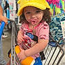 toddler, child, shopping_cart, hat, yellow_hat, pacifier, pink_clothing, curly_hair, store, clothing_rack, colorful_clothes, orange_cup, baby, footwear, person, retail, shopping, cute, indoor, casual