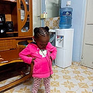 Elianah is registered to the contest to win money with this photo: braided_hair, child, clothing, door, floor, floor_tiles, furniture, girl, home, indoor, mirror, patterned_leggings, pink_hoodie, portrait, sink, socks, standing, water_dispenser, wooden_cabinet, young