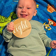 Owen is registered to the contest to win money with this photo: baby, blanket, cartoon, child, close_up, colorful, eight_months, face, family_photo, green_clothing, hands, happy, indoor, infant, lying_down, milestone, portrait, smiling, tongue_out, wooden_sign