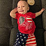 american_flag, baby, celebration, child, clothing, couch, cute, face, happy, indoor, infant, laying_down, person, plush, portrait, red_shirt, shorts, six_months, smiling, wooden_plaque