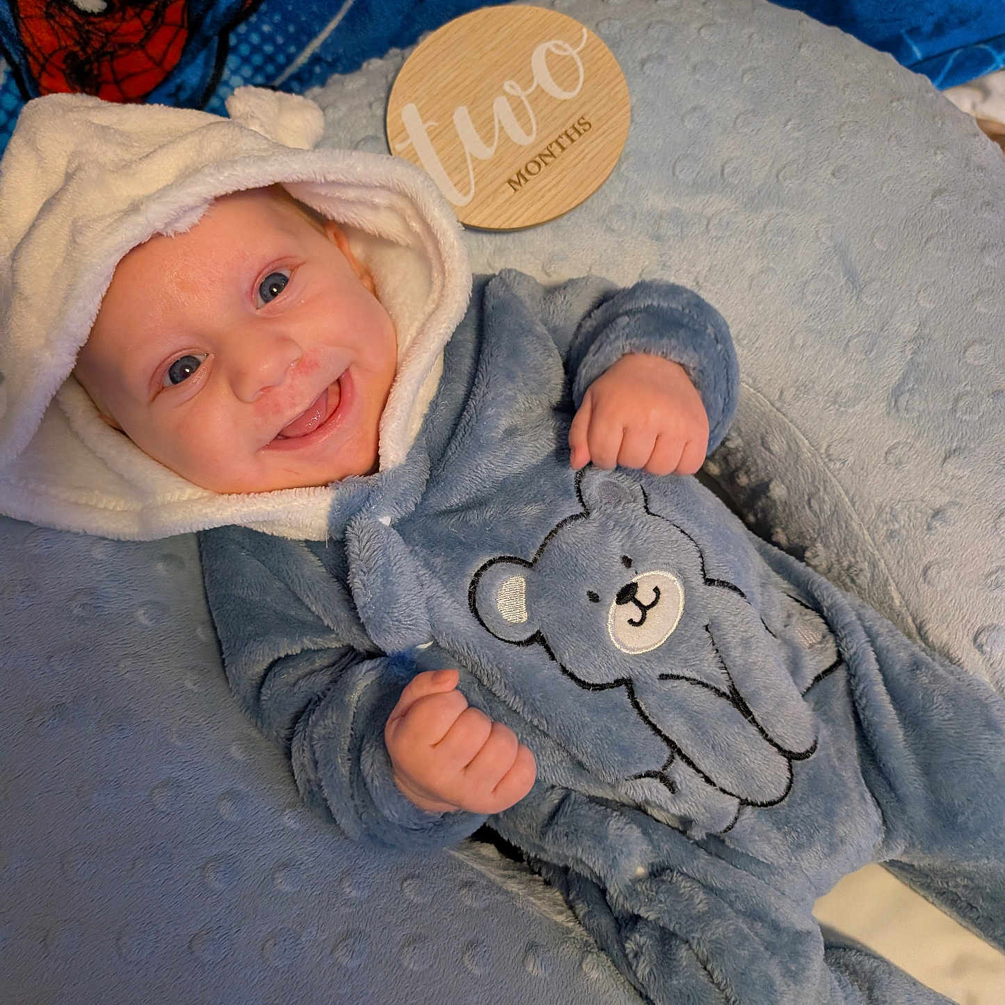 Owen is registered to the contest to win money with this photo: baby, smiling, blue_onesie, bear_design, hood, cushion, soft_texture, spiderman_blanket, wooden_plaque, two_months, infant, happy, cute, lying_down, child, portrait, indoors, cozy, apparel, face