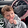 baby, child, car_interior, steering_wheel, pajamas, grey_clothing, hand, face, chewing, happy, person, seat, window, rain, car_mirror, storefront, pharmacy, green_shirt, pink_steering_wheel_cover, daytime