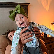 Marley joined the competition — help win amazing prizes! baby, child, infant, smiling, laughing, bow, headband, outfit, overalls, animal_print, held, adult, parent, hands, indoor, bedroom, bed, lamp, warm_lighting, portrait