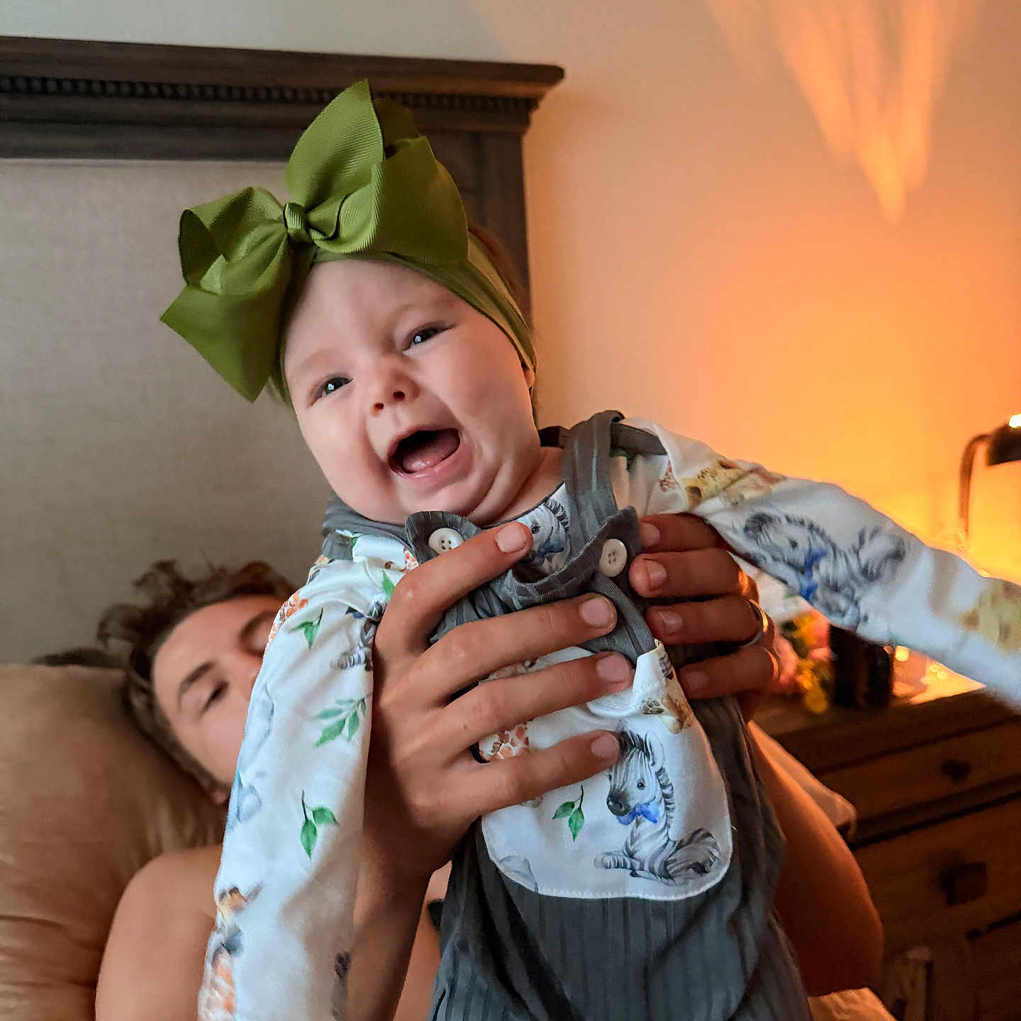 Marley joined the competition — help win amazing prizes! baby, child, infant, smiling, laughing, bow, headband, outfit, overalls, animal_print, held, adult, parent, hands, indoor, bedroom, bed, lamp, warm_lighting, portrait