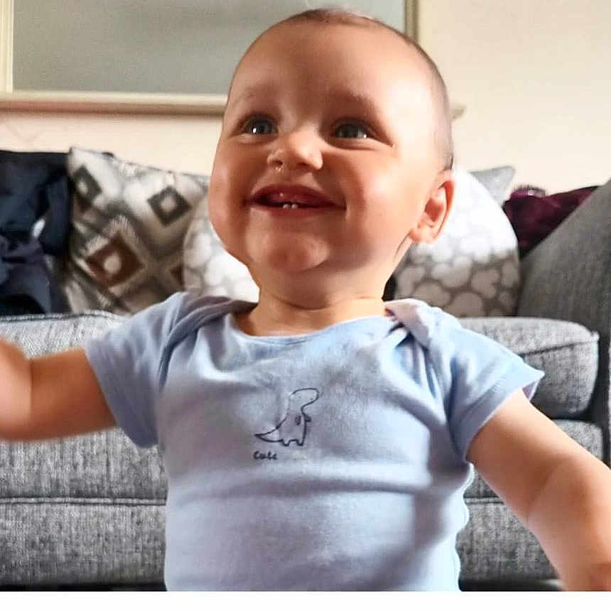 Noah joined the competition — help win amazing prizes! baby, blue_shirt, child, couch, cute, dinosaur_print, face, furniture, happy, home, indoor, infant, person, pillow, portrait, sitting, smiling, teeth, toddler, young_child