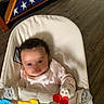 baby, infant, baby_seat, toy, mittens, curly_hair, pink_clothing, wooden_floor, flag_display, american_flag, triangular_flag_case, indoor, child, person, cushion, smiling, cute, young_child, home, playtime
