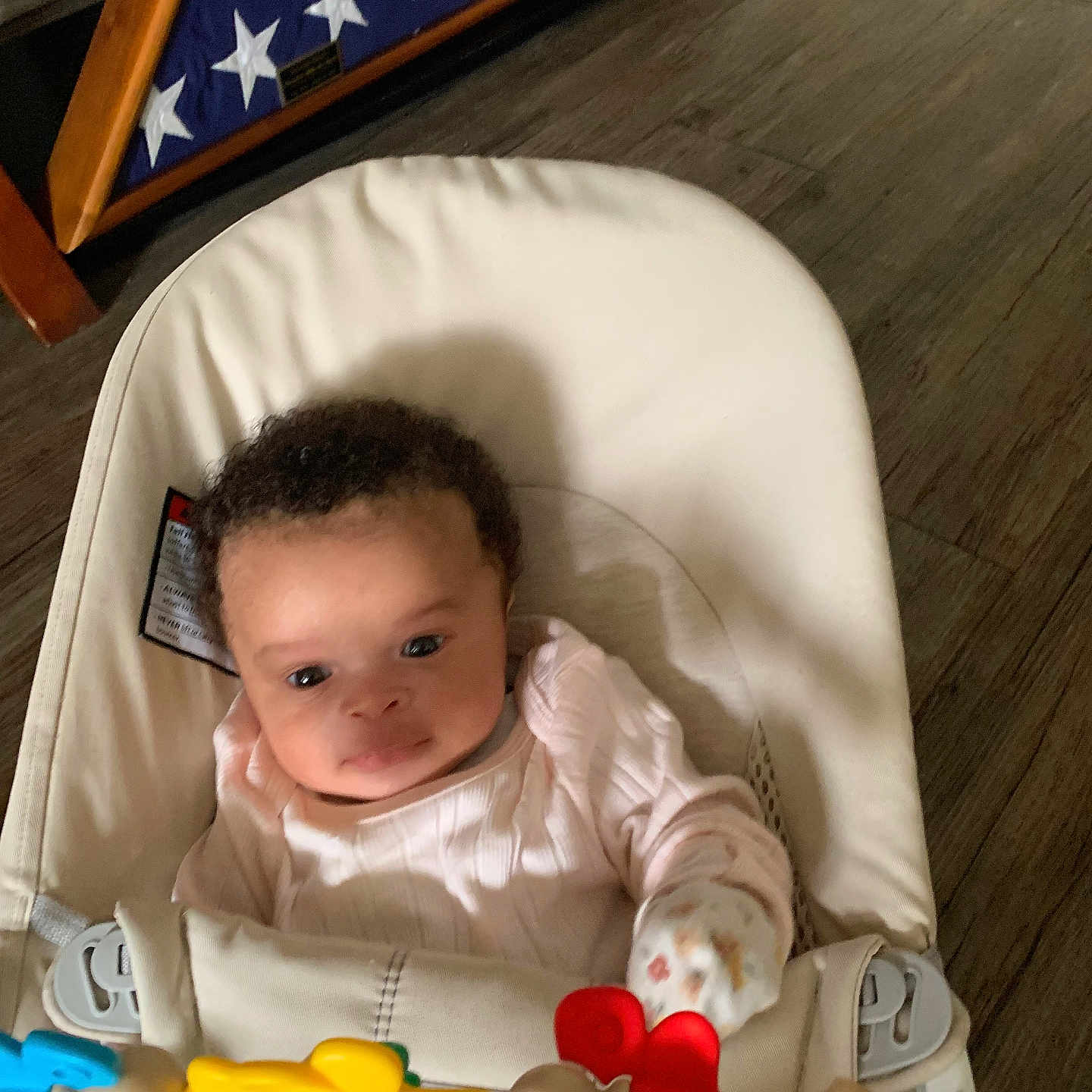 Amiylah joined the competition — help win amazing prizes! baby, infant, baby_seat, toy, mittens, curly_hair, pink_clothing, wooden_floor, flag_display, american_flag, triangular_flag_case, indoor, child, person, cushion, smiling, cute, young_child, home, playtime