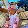 baby, infant, pink_clothing, headband, bow, bed, blankets, striped_bedding, tv_screen, cartoon, charging_cable, person, indoor, cute, child, sitting, expression, hand, foot, furniture