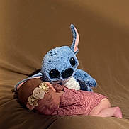 Emy a rejoint le concours — aidez-le/la à gagner de superbes lots ! newborn, baby, sleeping, plush_toy, stitch, flower_headband, pink_lace, soft_blanket, brown_background, cute, adorable, infant, resting, toy, child, portrait, indoor, cozy, peaceful, soft_lighting