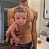 baby, child, person, man, shirtless, bathroom, mirror, sink, toiletries, toothbrush, hand, curly_hair, flooring, tile_wall, mirror_reflection, container, grooming, expression, holding, indoor
