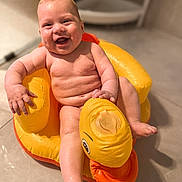 Cedric is registered to the contest to win money with this photo: baby, child, infant, smiling, happy, water, wet_skin, inflatable, duck, yellow, orange, bathroom, floor, toy, seat, sitting, cute, skin, face, joyful