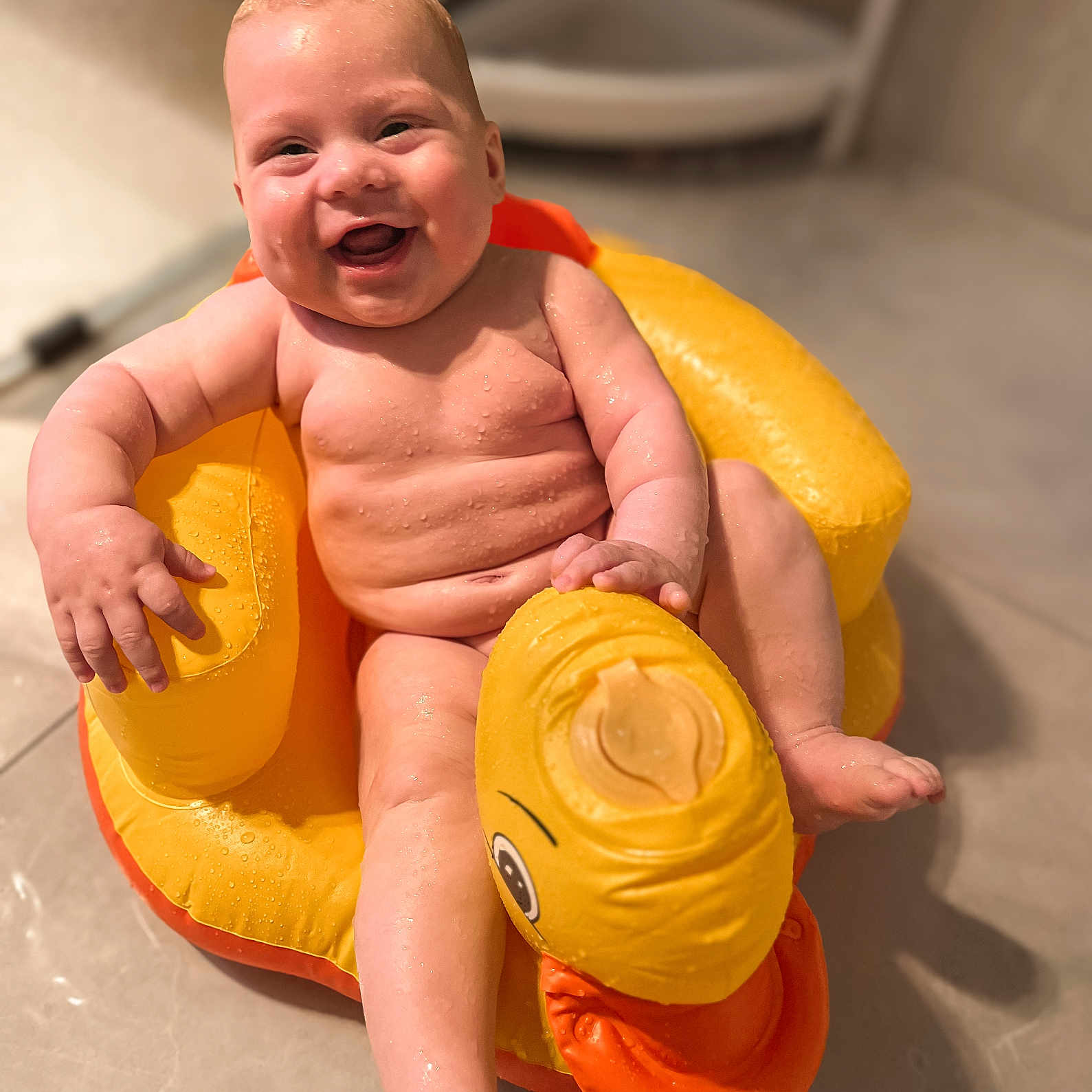 Cedric is registered to the contest to win money with this photo: baby, bathroom, child, cute, duck, face, floor, happy, infant, inflatable, joyful, orange, seat, sitting, skin, smiling, toy, water, wet_skin, yellow
