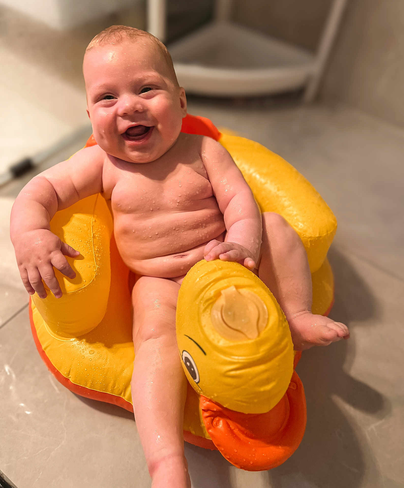 Cedric is registered to the contest to win money with this photo: baby, child, infant, smiling, happy, water, wet_skin, inflatable, duck, yellow, orange, bathroom, floor, toy, seat, sitting, cute, skin, face, joyful