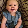 baby, child, smiling, blue_dress, sitting, couch, patterned_cushion, happy, indoor, cute, infant, footwear, small_hands, eyes, face, skin, cheeks, short_hair, buttons, soft_lighting
